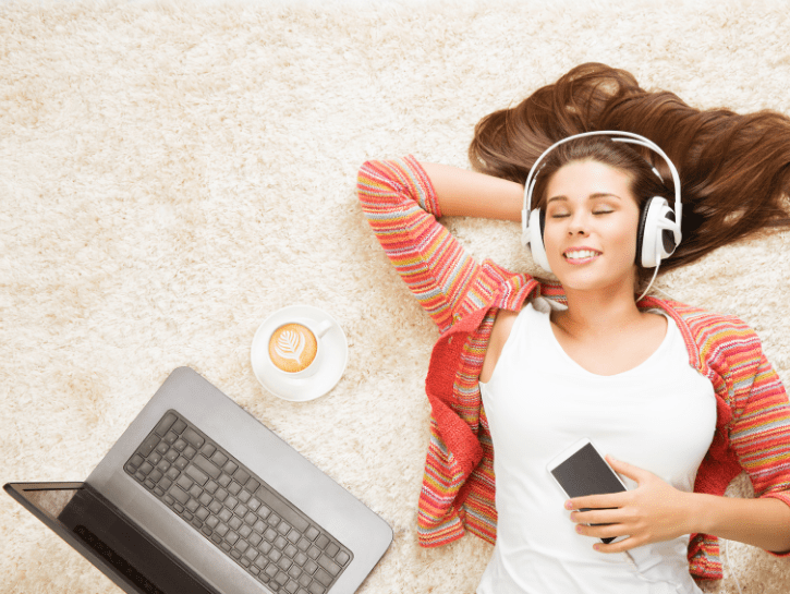 A woman laying down with headphones, holding a phone, next to a laptop and cup of coffee