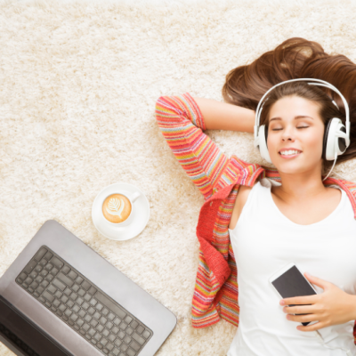 A woman laying down with headphones, holding a phone, next to a laptop and cup of coffee