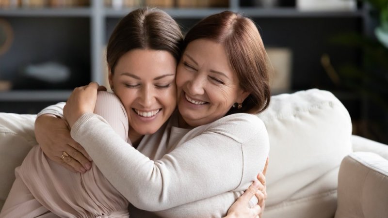 Affectionate loving middle-aged mother cuddling grownup child, amazed by good news. Excited to see daughter, happy senior older mom cuddling her, sitting together on a comfortable sofa in the living r