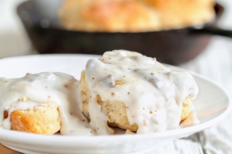 American biscuits from scratch covered with thick white sausage gravy. Selective focus with cast iron skillet/pan in the background over a white table.