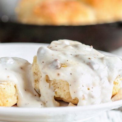 American biscuits from scratch covered with thick white sausage gravy. Selective focus with cast iron skillet/pan in the background over a white table.