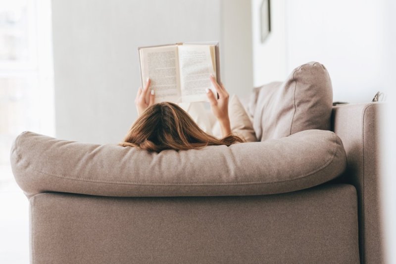 Back view of woman lying on sofa and reading book at home