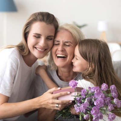 Cheerful feminine three diverse generations indoors. Sweet granddaughter mom and laughing grandmother sitting on couch in living room hold gift box and flowers embracing celebrating mother day at home