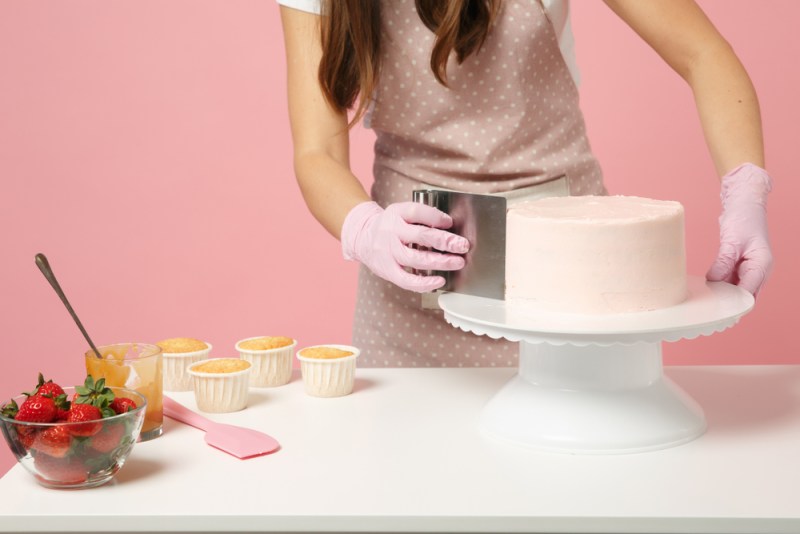 Close up cropped chef cook confectioner or baker in white t-shirt cooking at table isolated on pink pastel background in studio. Cream application cake making process. Mock up copy space food concept