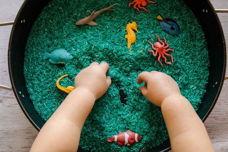 Close up of toddler hands playing with the blue rice in an ocean theme sensory tray play set up for toddler to explore