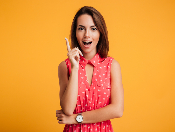 Close-up portrait of charming young woman in red dress pointing with finger, looking at camera, isolated on yellow background