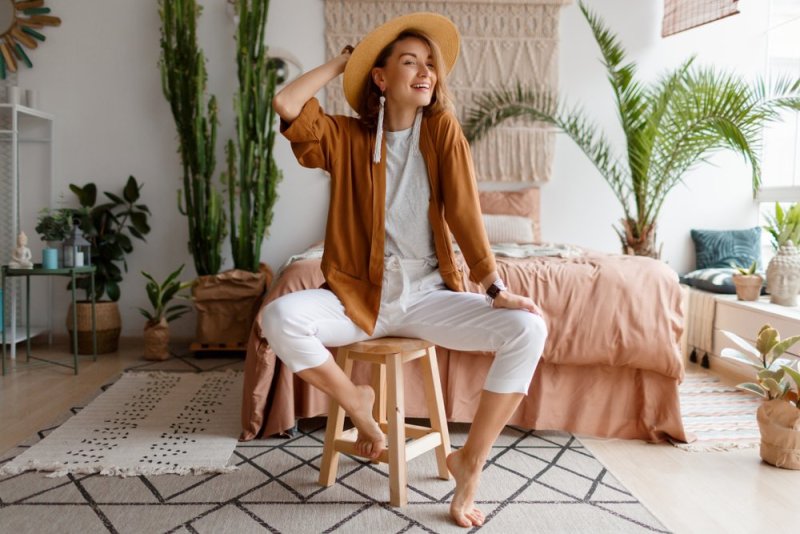 Cozy home atmosphere. Stylish woman in linen clothes sitting on chair over bed and home plants. Soft colors.