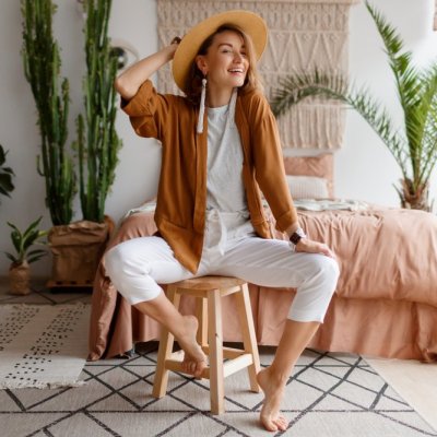 Cozy home atmosphere. Stylish woman in linen clothes sitting on chair over bed and home plants. Soft colors.
