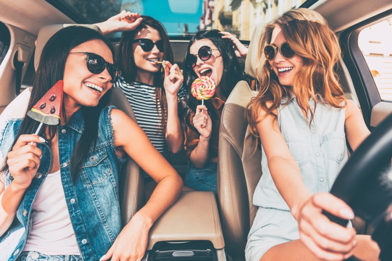 Great start of their journey. Four beautiful young cheerful women looking at each other with smile and holding lollipops while sitting in car