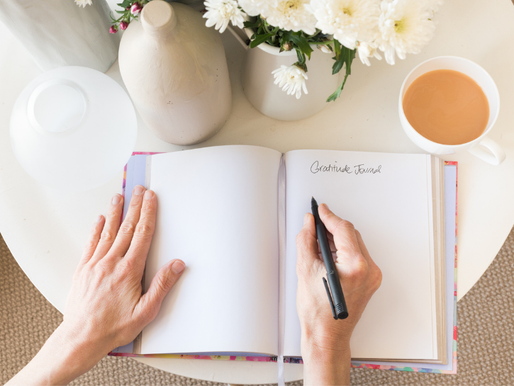 Hand holding pen writing in gratitude journal next to cup of coffee and flower vase