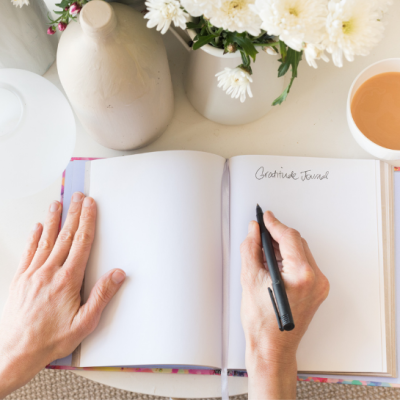Hand holding pen writing in gratitude journal next to cup of coffee and flower vase