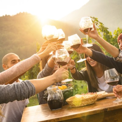 Happy friends having fun outdoor - Young people enjoying harvest time together outside at farm house vineyard countryside - Youth friendship concept - Hands toasting red wine glass at winery on sunset