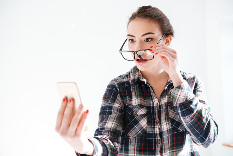 Image of confused woman wearing glasses standing over white background while look at phone