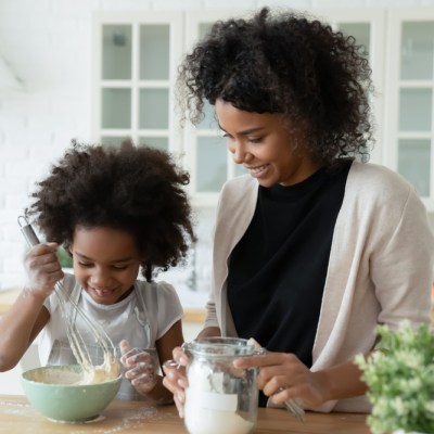 Little daughter helper and African mother cooking together in kitchen, kid girl preparing cake or pancakes enjoy process mixes dough using whisk, teach and upbringing, healthy home food, hobby concept