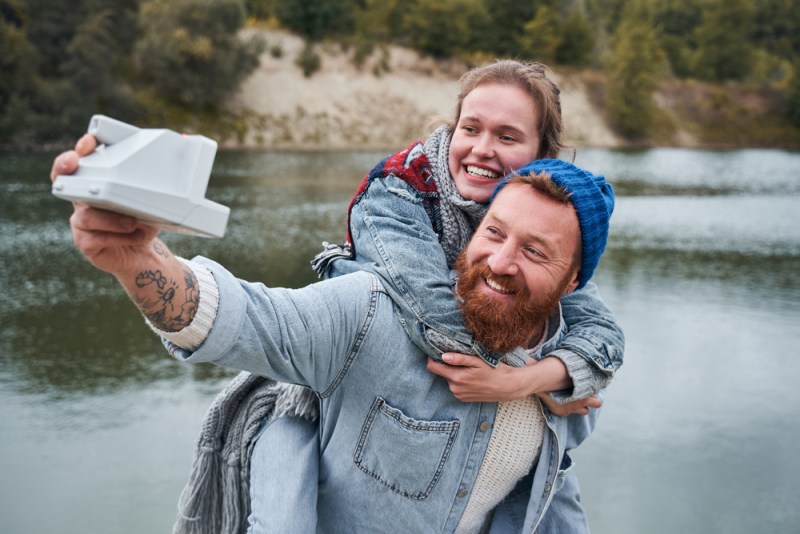 Love and date concept. Young happy caucasian woman brightly smiling while her beloved male carefully holding her on his back. Couple taking selfie with a Polaroid