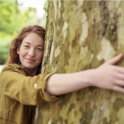 Nature-Lover Blond Teen Girl Hugging Huge Tree Trunk at the Park While Smiling at the Camera