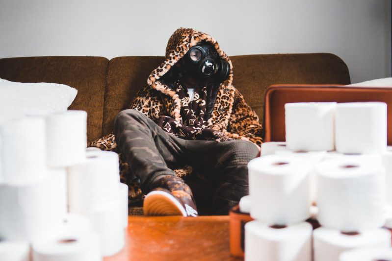 person with animal print hooded jacket sitting on couch with gas mask and toilet paper