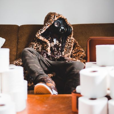 person with animal print hooded jacket sitting on couch with gas mask and toilet paper