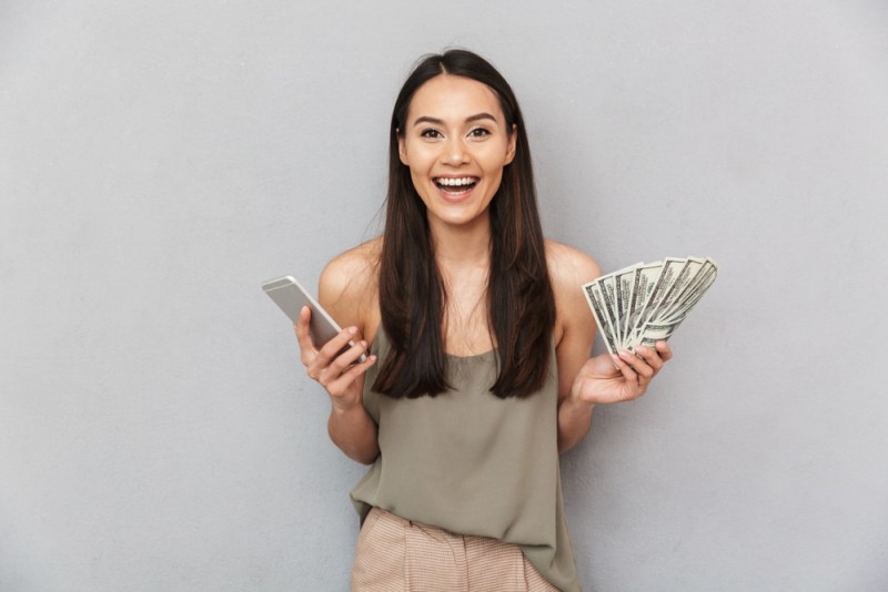 Portrait of an excited asian woman showing money banknotes and holding mobile phone isolated over gray background