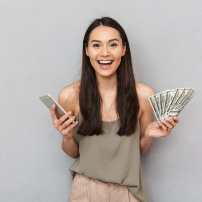 Portrait of an excited asian woman showing money banknotes and holding mobile phone isolated over gray background