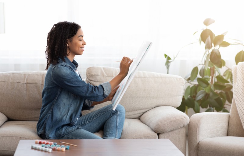 Quarantine Leisure. Young African American Woman Drawing Painting On Canvas At Home, Sitting On Couch In Living Room, Side View