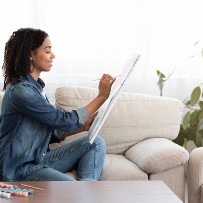 Quarantine Leisure. Young African American Woman Drawing Painting On Canvas At Home, Sitting On Couch In Living Room, Side View