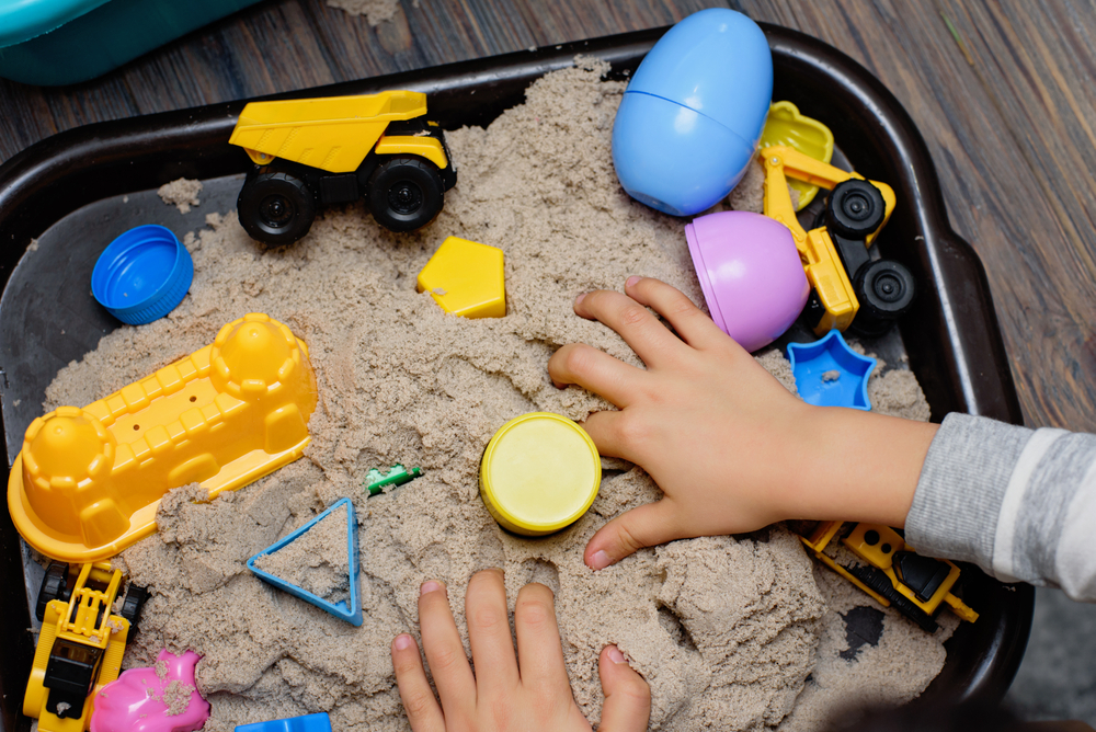 Sensory bin for sensory play. Child playing with kinetic sand and toy construction machinery. Hand of child in sand close up. Activity for fine motor development