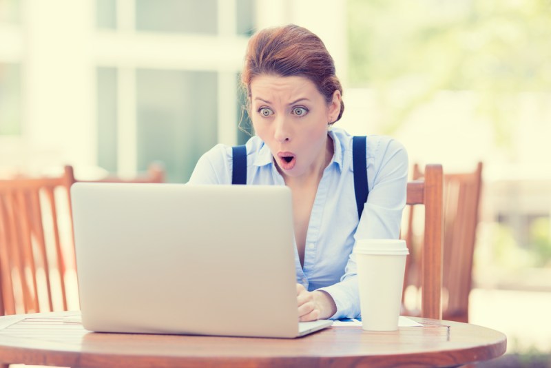Shocked young businesswoman using laptop looking at computer screen blown away in stupor sitting outside a corporate office. Human face expression, emotion, feeling, perception, body language, reactio