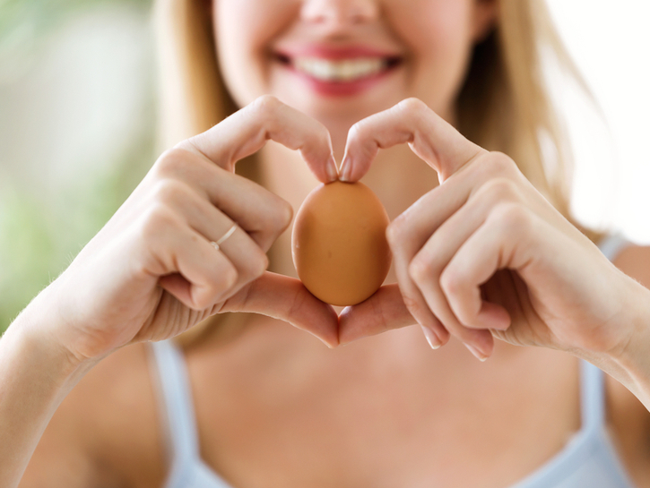 Shot of beautiful young woman showing brown chicken egg with hands in a heart shape at home.
