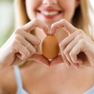 Shot of beautiful young woman showing brown chicken egg with hands in a heart shape at home.