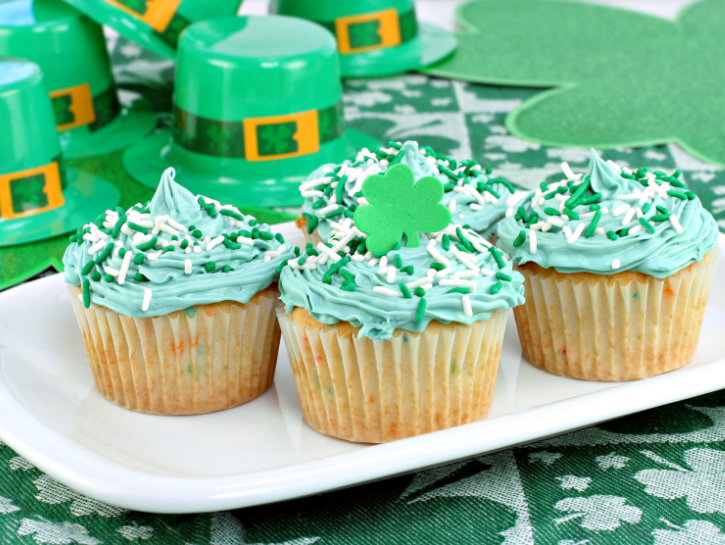 St. Patrick's Day themed cupcakes on white plate in front of plastic leprechaun hats