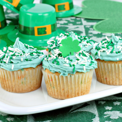 St. Patrick's Day themed cupcakes on white plate in front of plastic leprechaun hats