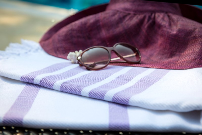 The concept of summer accessories close-up of white and purple Turkish towel, sunglasses and straw hat on rattan lounger with a blue swimming pool as background. Selective focus on the towel.