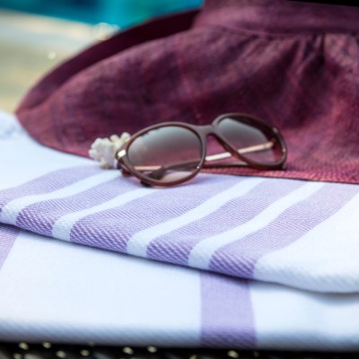 The concept of summer accessories close-up of white and purple Turkish towel, sunglasses and straw hat on rattan lounger with a blue swimming pool as background. Selective focus on the towel.