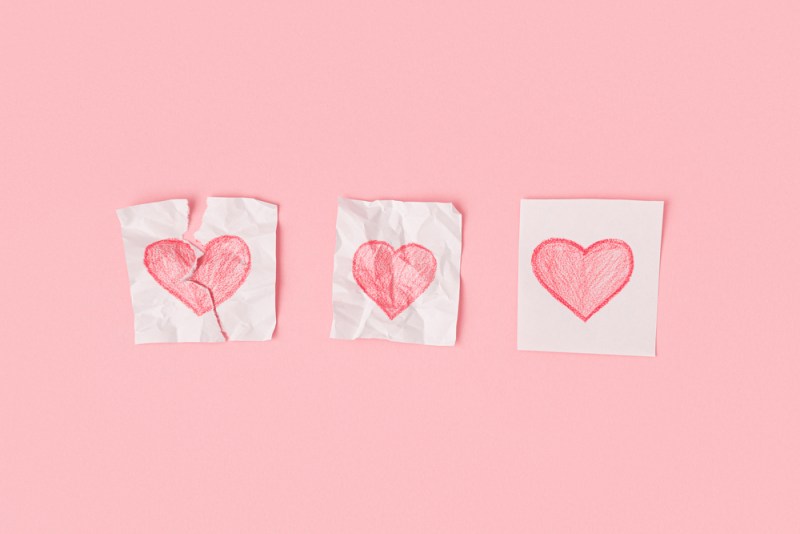 Three hearts drawn on pieces of paper on a pink background. One heart is torn, the second is crumpled, the third is whole. Progress of love.