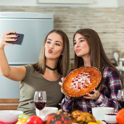 Two young women are taking selfies with their smart phone. They are taking pictures of food they made for Thanksgiving dinner.