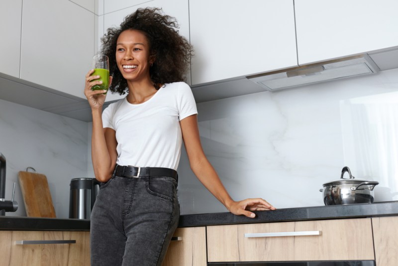 Woman drinking green detox juice, smoothie drink in kitchen portrait. Happy smiling african girl with glass of healthy fresh raw vegetable smoothie at home. Diet nutrition concept