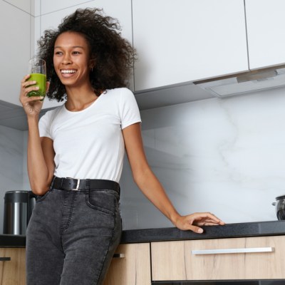 Woman drinking green detox juice, smoothie drink in kitchen portrait. Happy smiling african girl with glass of healthy fresh raw vegetable smoothie at home. Diet nutrition concept