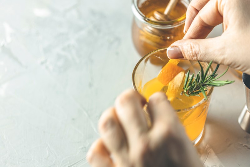 Woman hand decorate orange twist honey bourbon cocktail with rosemary simple syrup or homemade whiskey sour cocktail drink