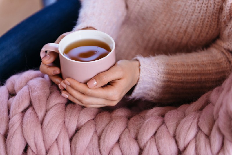Woman holding cup of tea wearing pink wool sweater with matching nails and Merino wool blanket