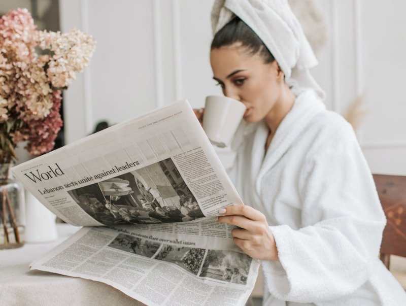 woman in white robe reading newspaper and drinking coffee