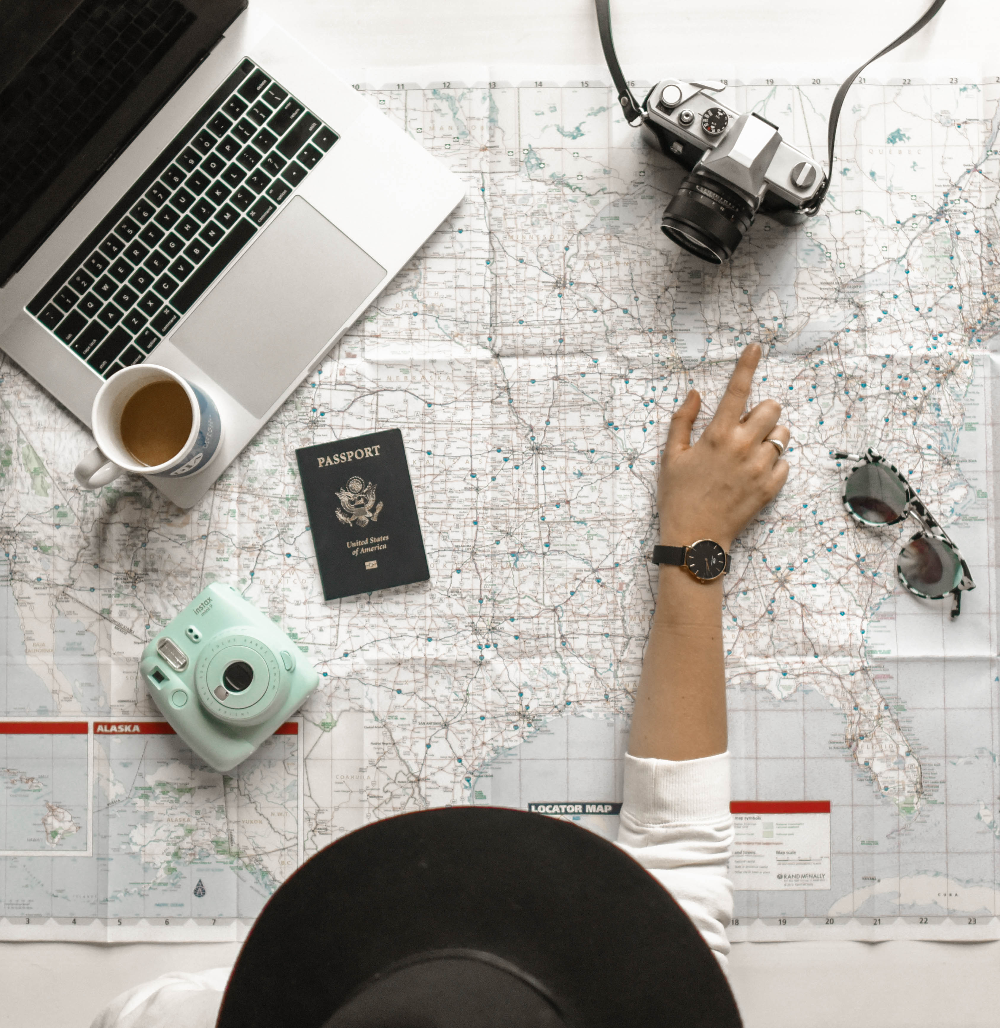 Woman wearing hat pointing at map of US with laptop, cameras, Passport, and coffee in background