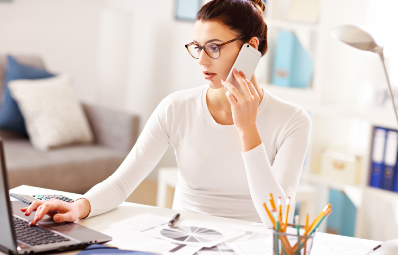 woman with glasses holding phone working on laptop in home office