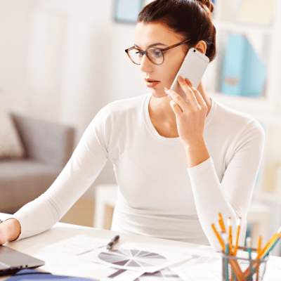 woman with glasses holding phone working on laptop in home office