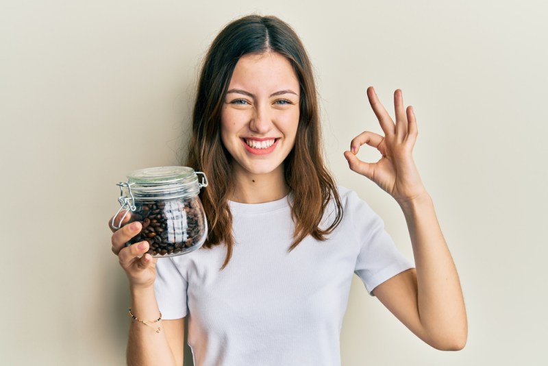 Young brunette woman holding jar with coffee beans doing ok sign with fingers, smiling friendly gesturing excellent symbol