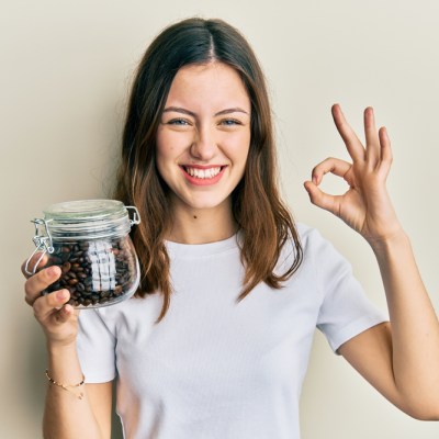 Young brunette woman holding jar with coffee beans doing ok sign with fingers, smiling friendly gesturing excellent symbol