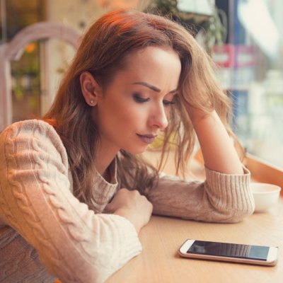 Young woman sitting at table in cafe looking at phone being unhappy with breakup.