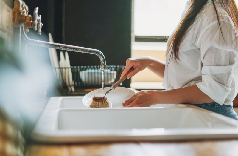 Young woman washes dishes with wooden brush with natural bristles at window in kitchen. Zero waste concept