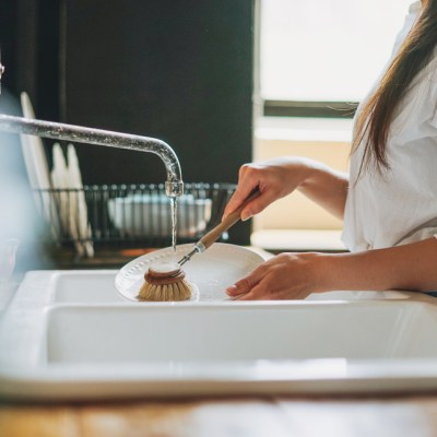 Young woman washes dishes with wooden brush with natural bristles at window in kitchen. Zero waste concept