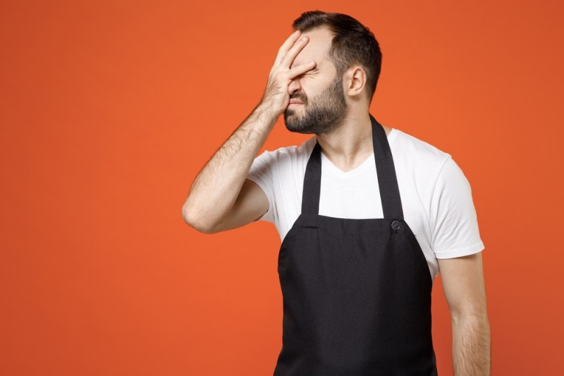 Young confused man barista bartender barman employee in black apron white t-shirt work in coffee shop put hand on face facepalm epic fail gesture isolated on orange background Small business startup